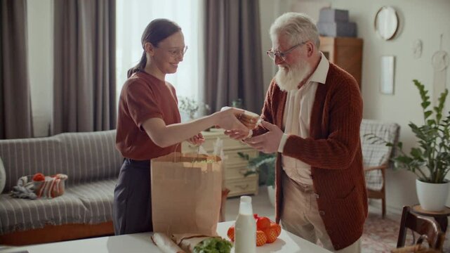 Young woman unpacking fresh groceries from paper bag for elderly grandfather at home, providing family support and helping with daily routine