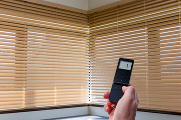 Wooden blinds automated, beige color, on large corner windows. Mans hand holds black remote control panel for electric wood blinds, connected to smart home.