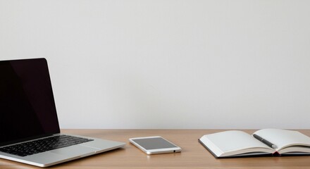 A workspace showing a laptop with a blank screen, a smartphone, and an open notebook. The items are neatly arranged on a wooden desk with a plain white wall in the background.