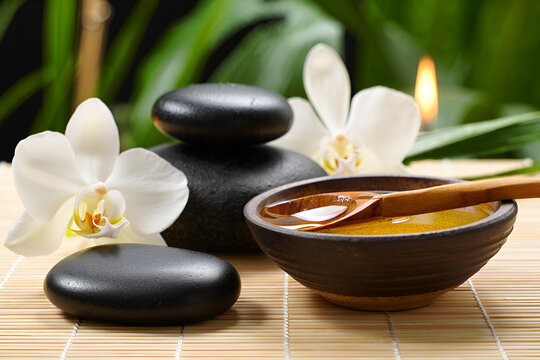 A serene spa still life composition featuring soft candlelight, smooth black massage stones, and fresh white orchids arranged on a bamboo mat. A small bowl of essential oil with a delicate wooden spoo