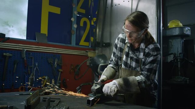 Female welder handling portable electric angle grinder to grind metal in a workshop, medium shot. Craft, work, and manufacturing concepts.