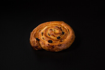 Puff pastry snail with maple syrup and raisins on a black background, top view