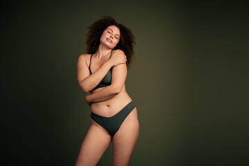 Fototapeta premium Confident young woman embracing her natural beauty in lingerie with a khaki green background, celebrating body positivity