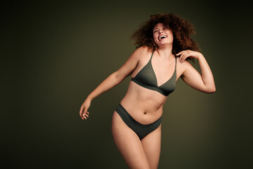 Fototapeta premium Confident young woman in green lingerie with curly brown hair showcasing natural beauty and self confidence on a khaki background
