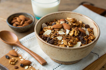 Fototapeta premium A rustic ceramic bowl with homemade low-carb granola, filled with roasted nuts, coconut flakes, cacao nibs. A wooden spoon rests on the side, and small glass of almond milk is slightly out of focus