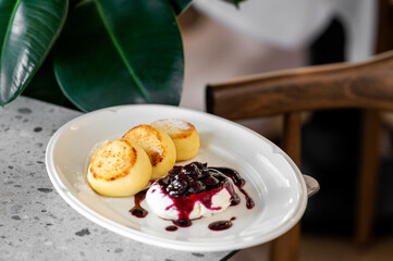 Three golden-brown cheese pancakes on a white plate, topped with berry sauce and a dollop of cream. The dish is placed on a speckled countertop with green leaves in the background.