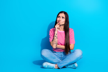 Thoughtful young woman in striped pink T-shirt sitting cross-legged with a contemplative expression...