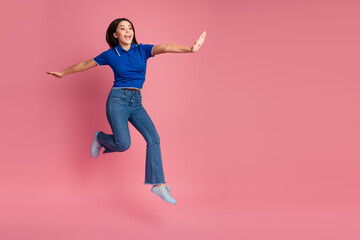 Young woman jumping joyfully with open arms in casual fashion outfit on a pink background, exuding...