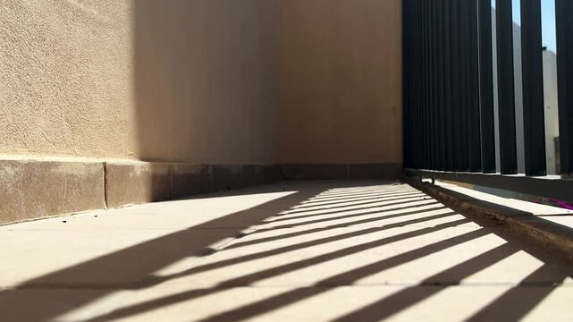 Sunlit outdoor corridor with strong geometric shadows from a metal fence. Warm beige walls and tiled flooring create a minimalist, modern aesthetic. Ideal for architectural and abstract concepts.