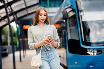 Young woman enjoying sunny day on urban street while using smartphone near public transport