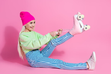 Joyful young woman in stylish casual outfit tying her roller skates against a vibrant pink background