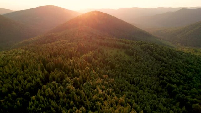 Aerial view of serene mountain landscape at sunset. Forested hills bathed in warm, golden light, creating tranquil atmosphere. Rolling hills stretch into the distance, covered in dense, lush greenery.