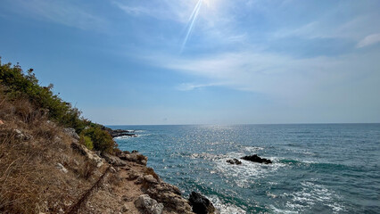 Rocky coastal path leading to the sparkling blue Mediterranean Sea in Alanya, Turkey, under a bright summer sun with clear skies and calm waves.