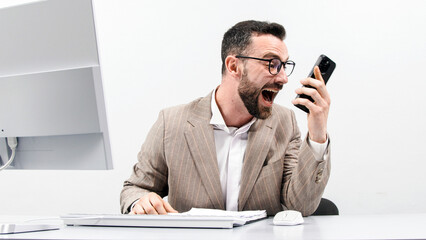 Angry businessman shouting and gesticulating while talking on cell phone in front of computer...