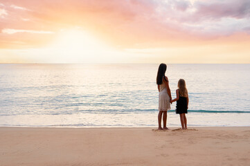 Mom and daughter holding hands on a beach, gazing at a colorful sunset over the ocean.