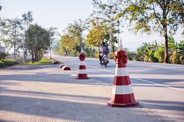 A row of red and white traffic cones placed along a road with trees and greenery in the background,...