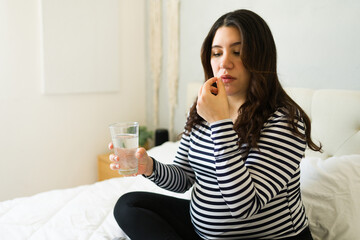 Pregnant woman seated on bed, swallowing prenatal vitamin with water, emphasizing medication management during pregnancy