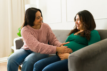 Happy pregnant woman feeling her baby kick while sitting on sofa and talking with doula during home...
