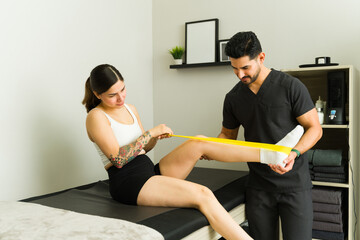 Latin woman doing leg physiotherapy exercises with resistance band assisted by professional therapist in his clinic