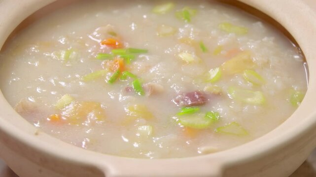 Deliciously steaming bowl of congee with vegetables, garnished with green onions, served in a traditional clay pot