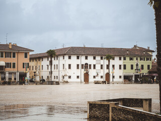 A rainy day in the historic town square of Palmanova, Italy, featuring old buildings with colorful facades, statues, and palm trees under a cloudy sky.