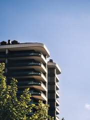 A modern high-rise residential building with elegant curved balconies against a clear blue sky. Sunlight casts shadows on the structure, while green tree tops in the foreground add contrast and depth.