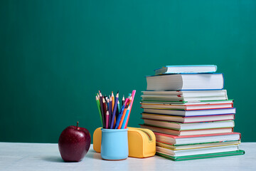 Stack of colorful books, a mug filled with colored pencils, an apple, and a yellow pencil case are...