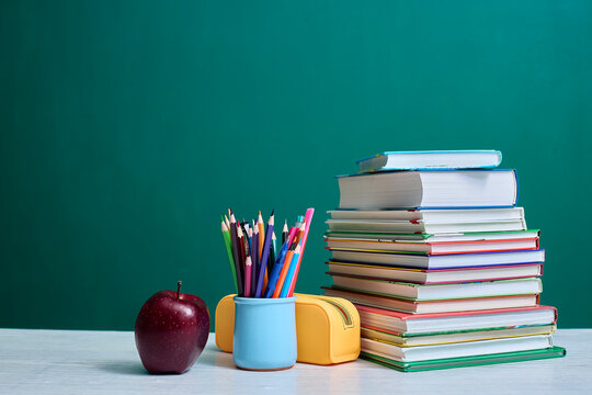 Stack of colorful books, a mug filled with colored pencils, an apple, and a yellow pencil case are arranged on a desk against a green chalkboard background, depicting academic theme.