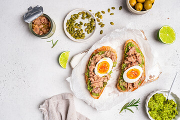 Tuna toast. Open sandwiches with whole grain bread, canned tuna, boiled egg, avocado and olives on white background. Healthy breakfast food, top view
