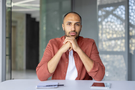A serious man looks directly at the camera while sitting at a desk, appearing to be on a video call.