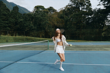 Obraz premium Joyful young female tennis player in white attire celebrating on a sunny outdoor court, showcasing athleticism and enthusiasm with a green nature backdrop