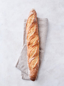 A long, crusty loaf of artisan bread rests on a soft linen cloth, inviting warmth and home-cooked comfort. Its golden-brown crust and intricate scoring showcase expert craftsmanship in baking