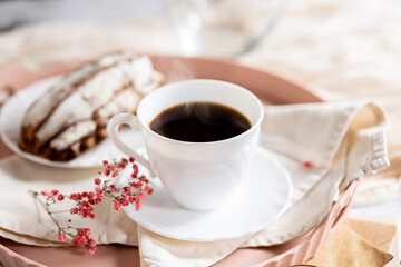 White cup filled with steaming coffee on a saucer, placed on a pink tray. Beside it lies a French eclair with cream. A small sprig of pink flowers adds decoration.