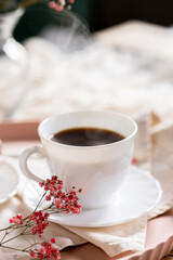 White cup filled with steaming coffee on a saucer, placed on a pink tray. Beside it lies a French eclair with cream. A small sprig of pink flowers adds decoration.