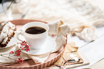 White cup filled with steaming coffee on a saucer, placed on a pink tray. Beside it lies a French eclair with cream. A small sprig of pink flowers adds decoration.