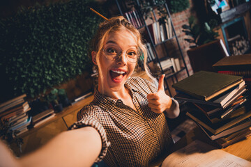 Energetic young woman studying indoors gives a thumbs up with a stack of books in a cozy home...