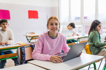 Smiling female student using a laptop in a classroom while classmates study in the background, showcasing technology in education