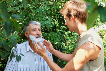 Obraz premium A young man helps an elderly man apply shaving foam on his face outdoors.