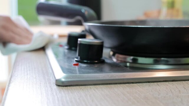 A hand wipes the stove surface near control knobs and a frying pan using a paper towel, showing kitchen cleanliness habits during cooking process