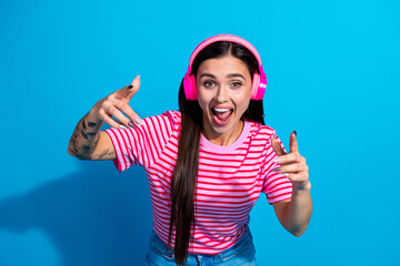 Smiling young woman with headphones gesturing lively in pink striped shirt against vibrant blue background, showcasing music enjoyment.