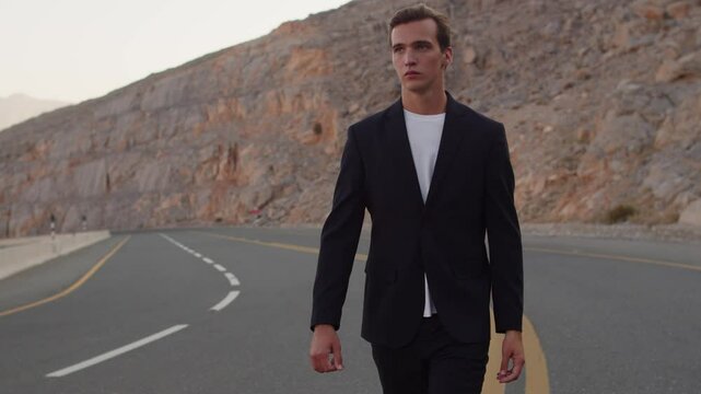  Confident young male model walks on road and looking camera. Handsome young guy model walking and turns around for camera. Male fashion model in a blue business suit looking back, on nature.