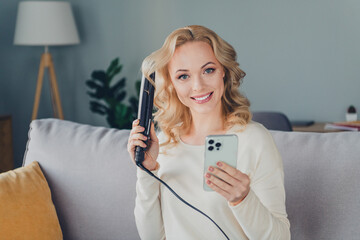Smiling blonde woman styling her hair with a curling iron at home, enjoying a comfortable weekend...