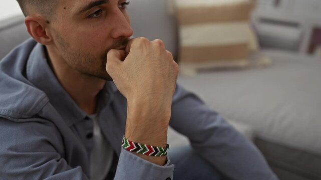 Young man sitting on a couch in a cozy living room, wearing a colorful bracelet, deep in thought.