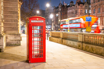 A classic red telephone box stands illuminated on a quiet street in London. Night falls as buses pass by, highlighting the city's vibrant nocturnal atmosphere.