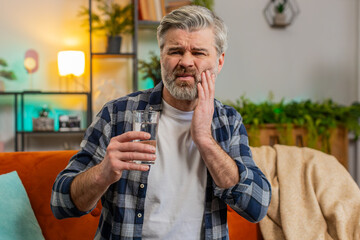 Senior bearded man feel terrible toothache after drink cold water. Middle aged guy sitting on sofa...