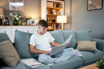 Obraz premium Focused student studying online and reviewing documents on sofa at home