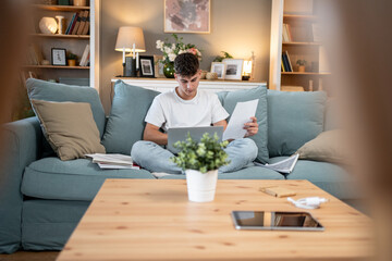 Focused student studying on sofa with laptop and documents at home