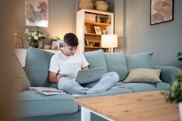 Focused teen studying on sofa with laptop and documents at home