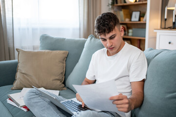 Focused teen studying on sofa with laptop and documents at home