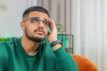 Portrait of sad Indian man guy sitting at home room looks pensive thinks over life concerns or...